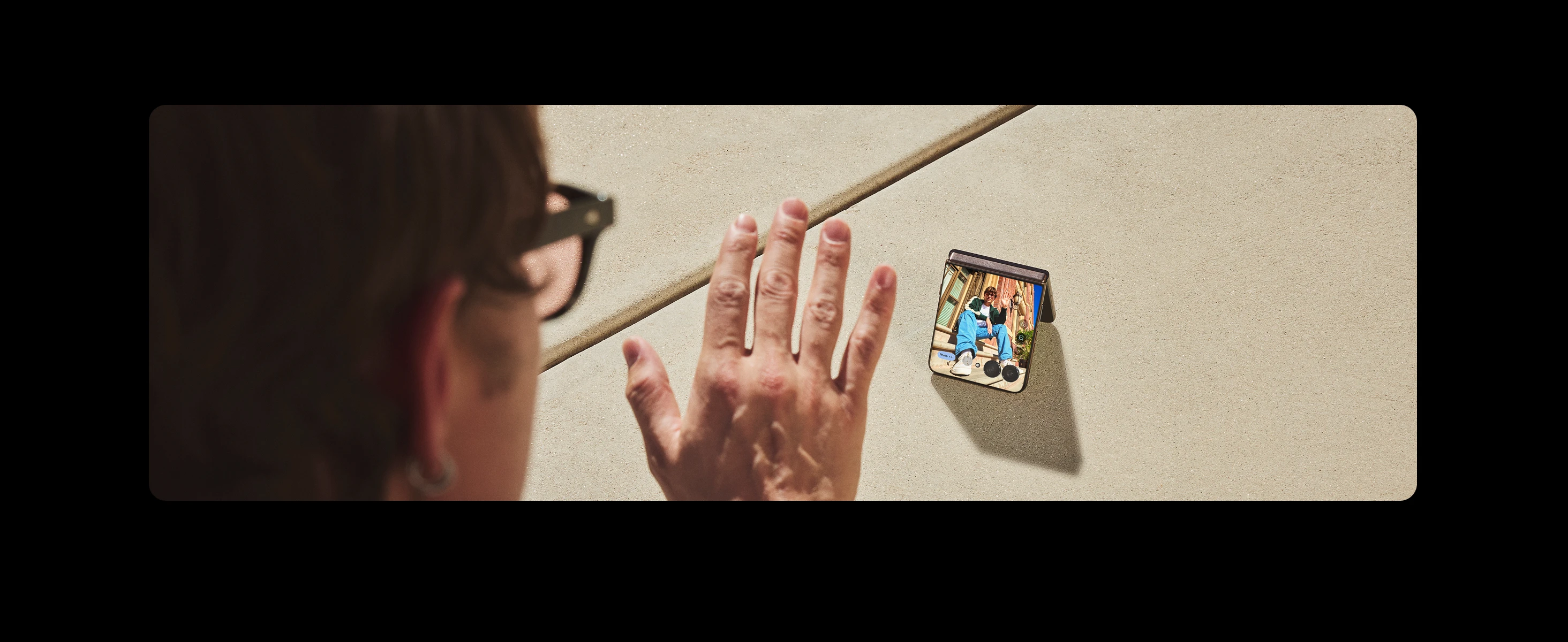 A person waves at a smartphone showing a video call with someone waving back, displayed on a beige concrete surface outdoors.