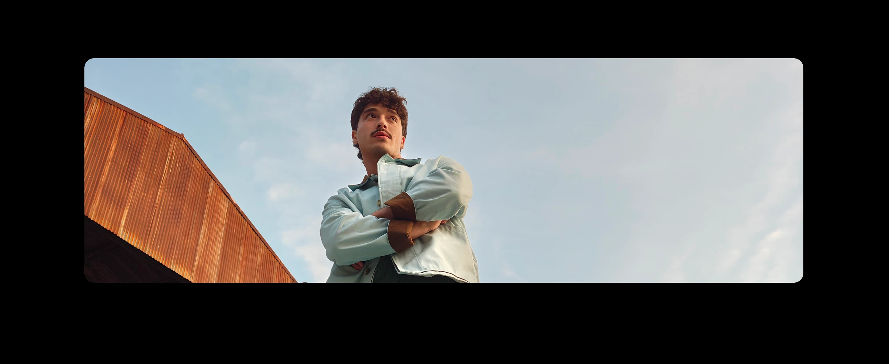 A young man with dark curly hair and a mustache looks up, arms crossed, standing next to a rust-coloured metal building under a blue sky.
