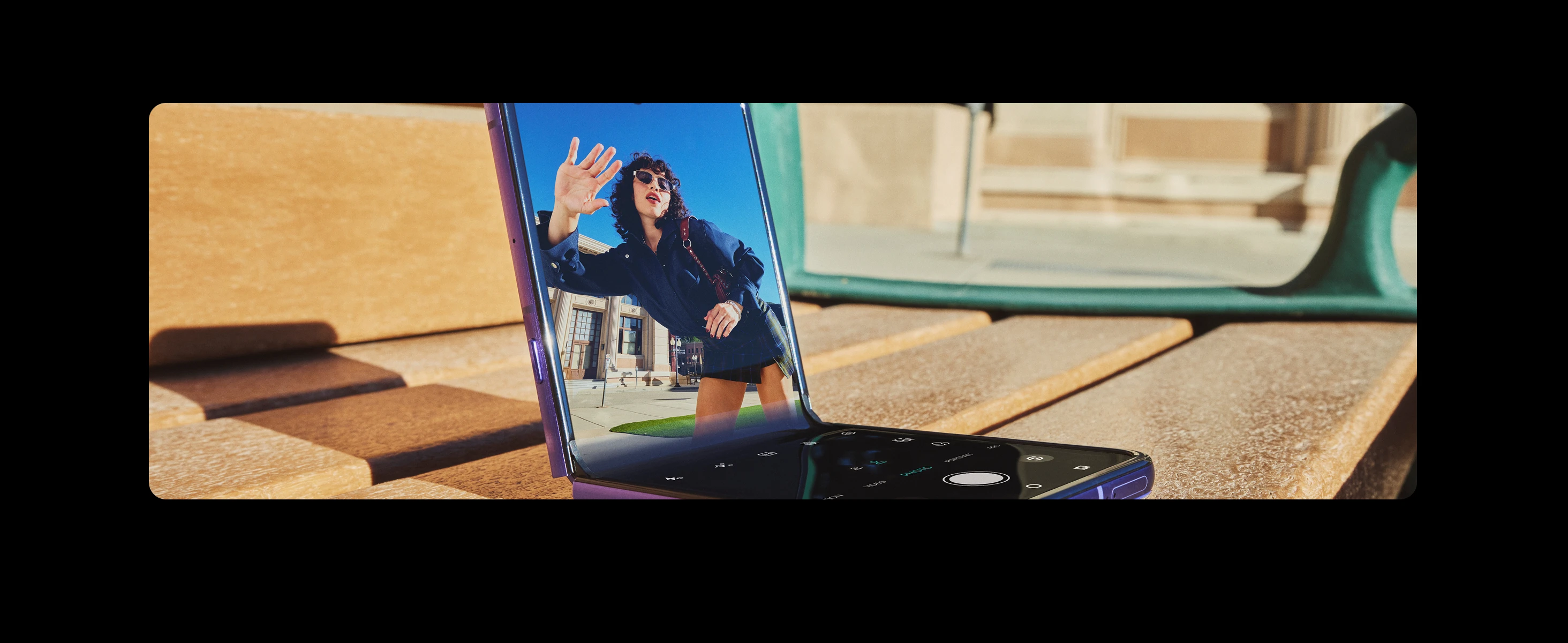 A foldable smartphone on a bench displaying a photo of a woman reaching out to the camera, with an outdoor urban setting and a building in the background.