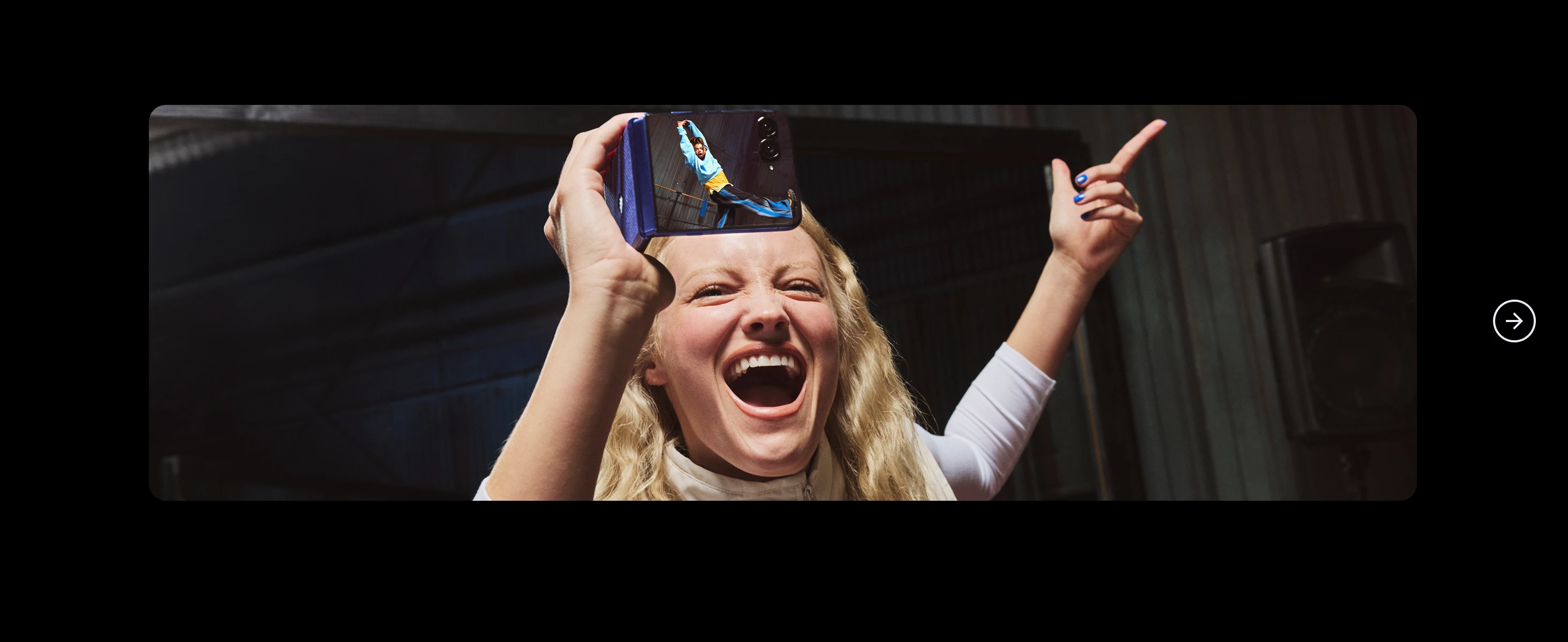 Smiling woman holds up a phone showing a man jumping, pointing upward excitedly in a dim industrial setting.