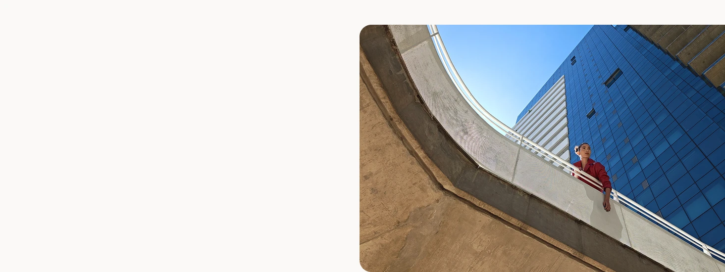 A low-angle outdoor shot of a person leaning on a curved concrete railing, with a tall glass building and clear blue sky in the background, demonstrating the phone’s wide-angle photography capability.