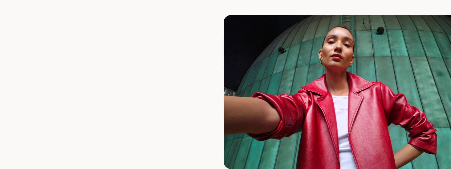 A close-up selfie-style image of a person wearing a bright red jacket, standing in front of a large green dome structure, showcasing the phone’s ultra-wide front camera perspective.