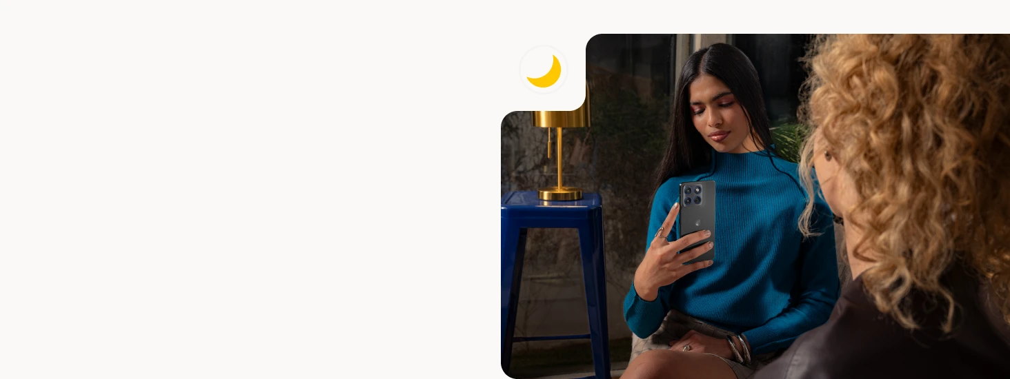 A person seated indoors holding a Motorola Edge 70 smartphone in one hand, with a lamp and another person partially visible in the background, suggesting a relaxed evening setting.