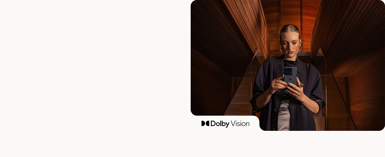 A person stands inside a warm wooden architectural interior holding a smartphone with both hands, framed by dramatic lighting and symmetrical paneling, with Dolby Vision branding displayed.