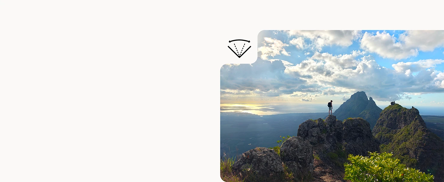 A wide scenic view of a person standing atop rocky terrain overlooking distant mountains, a valley, and reflective water under a cloud‑filled sky.