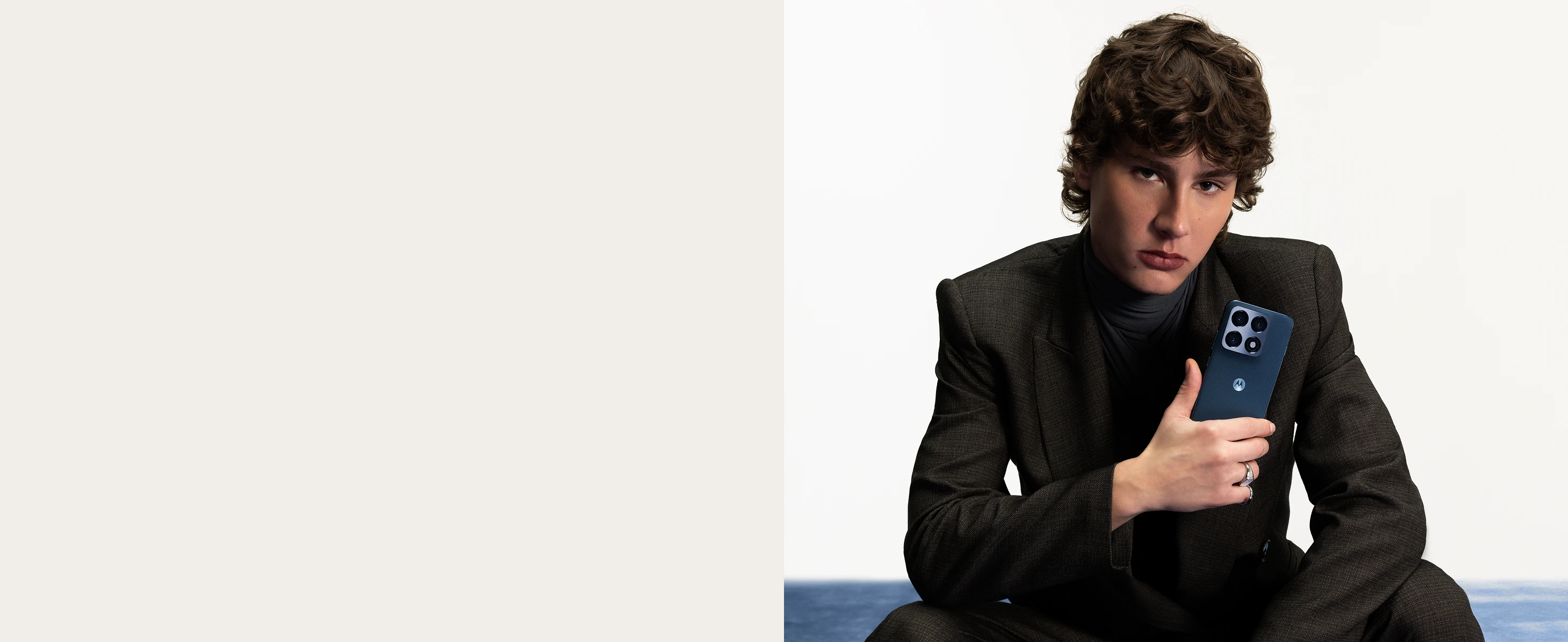 Young person with curly hair in a dark blazer holds a Motorola smartphone with triple cameras against a plain background.