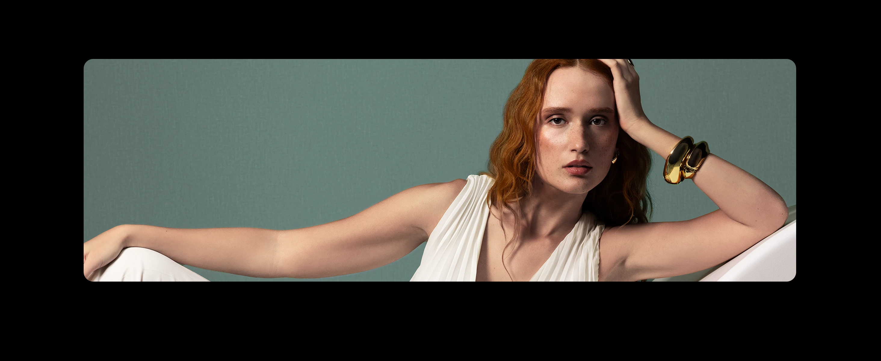 Portrait of a red-haired woman wearing white and gold bracelets, leaning her head on her hand against a plain teal background.