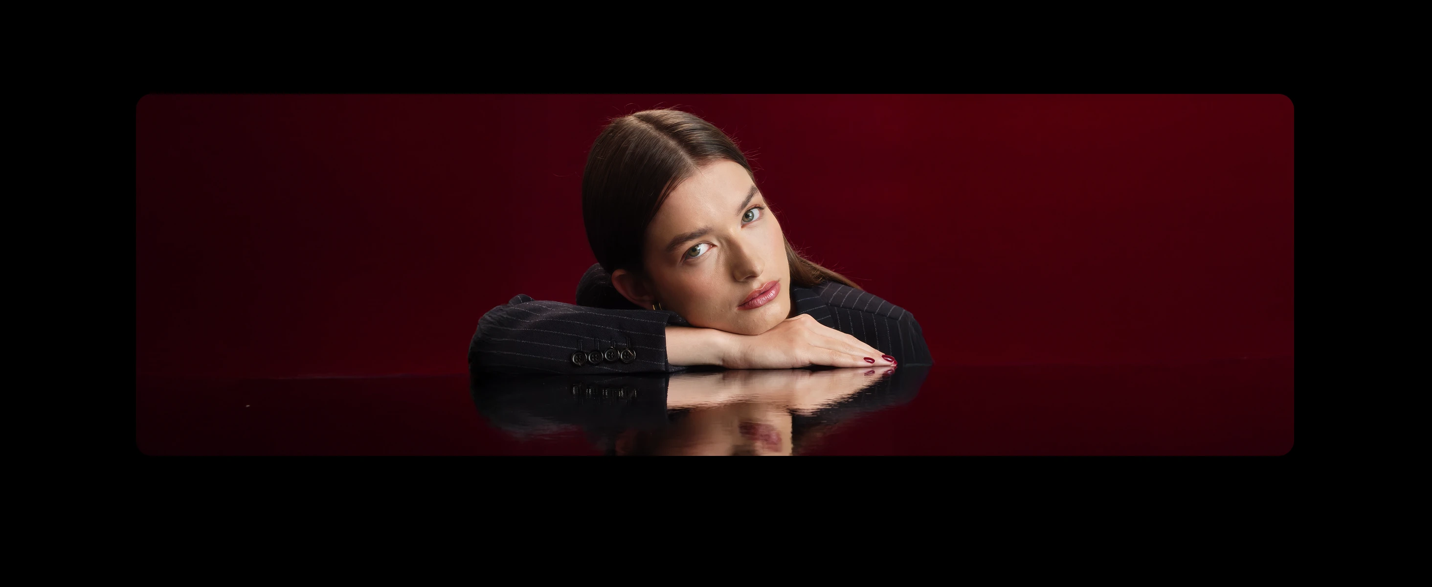 Portrait of a young woman resting her head on crossed arms on a reflective surface, set against a deep red background, with a neutral expression and dark pinstriped blazer.