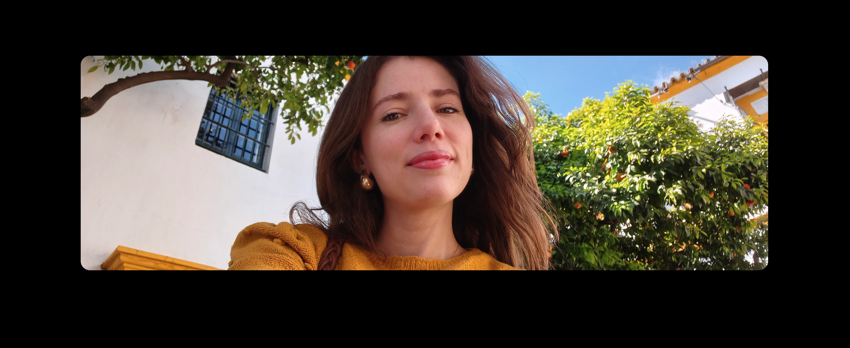 Selfie of a young woman in a mustard yellow sweater with an orange tree and white building behind her, under a clear blue sky.