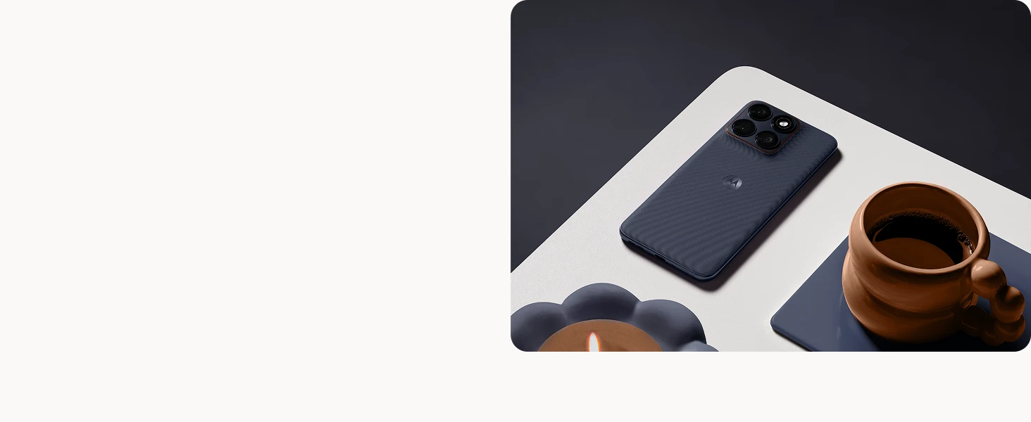 A dark blue Motorola smartphone lies face down on a white table beside a brown coffee cup on a blue coaster and a lit candle in a flower-shaped holder.