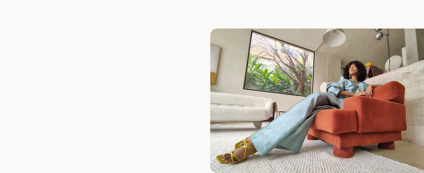A woman relaxes on an orange armchair in a bright, minimalist living room with a large window showing greenery outside.