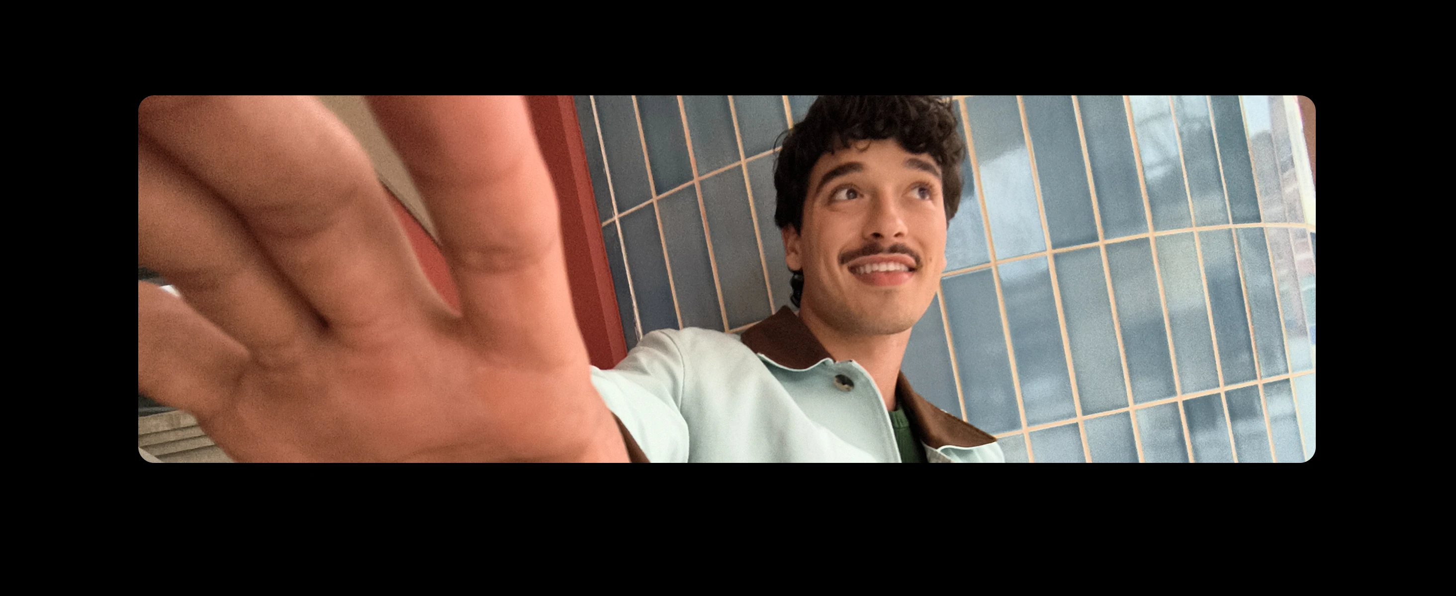 A young man with curly dark hair and a moustache smiles and reaches towards the camera, standing in front of a tiled wall.