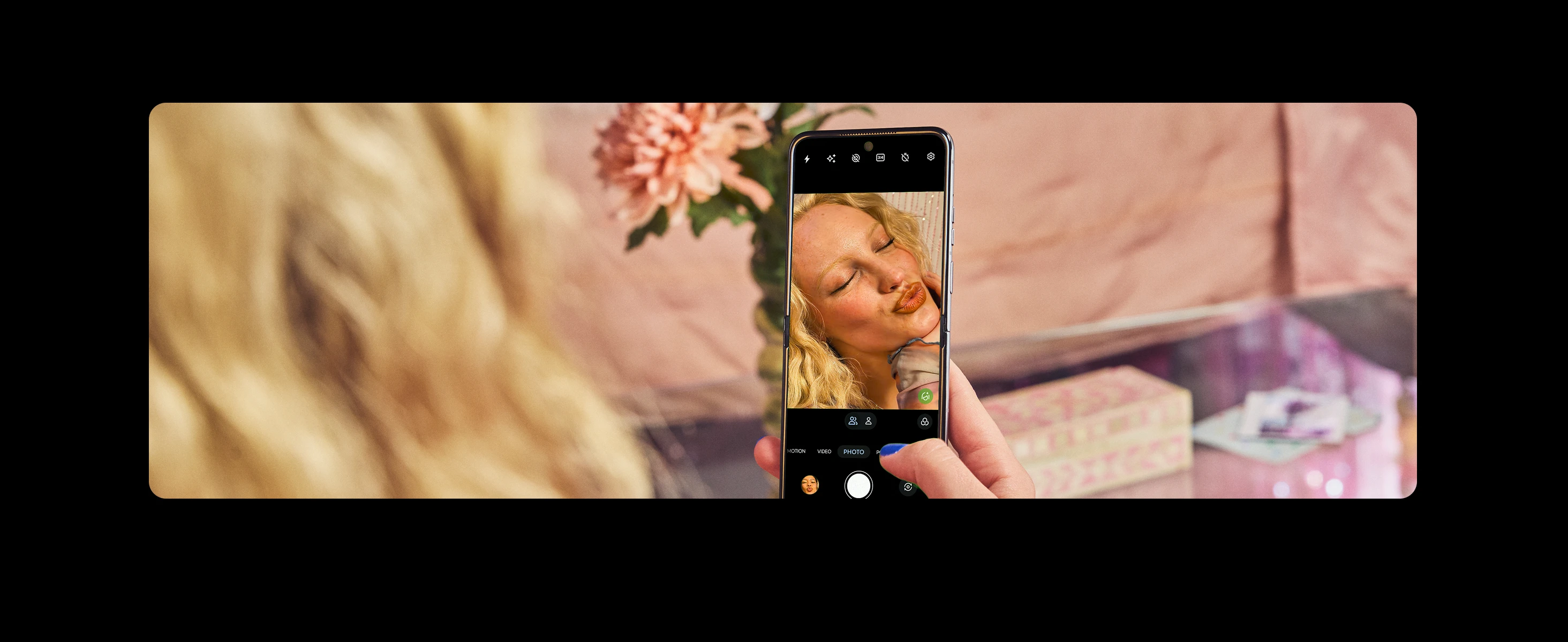 A blonde woman takes a selfie on her smartphone, puckering her lips, with a pink flower and table in the background.
