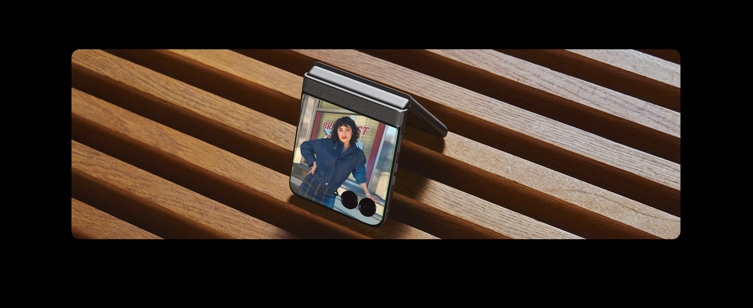 A foldable smartphone on a wooden slatted surface shows a woman in a blue blouse and plaid skirt posing with one hand on her hip. The background includes a sign with red and white text.