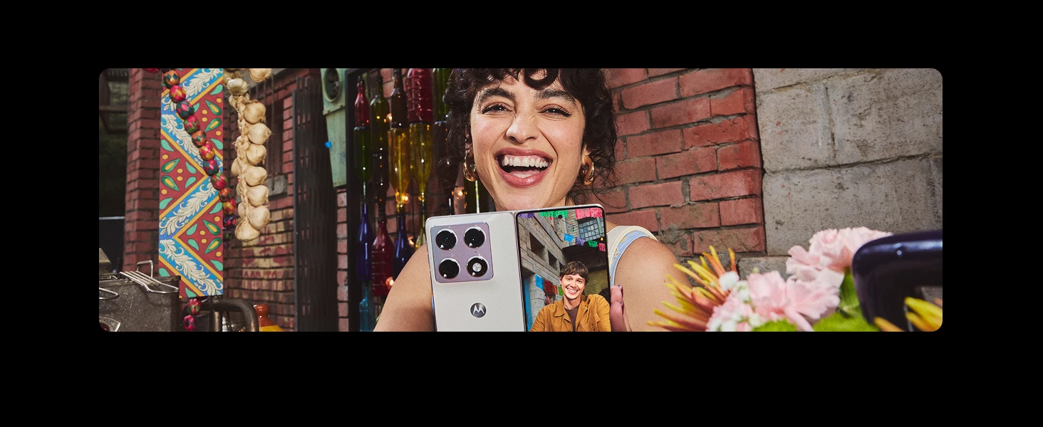 A happy woman holding a partially unfolded Motorola foldable phone showing a smiling man on a video call, against a brick wall decorated with hanging bottles and garlands.