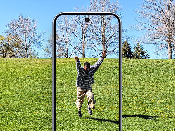 A man joyfully jumps in a grassy park, viewed through the frame of a Pixel phone